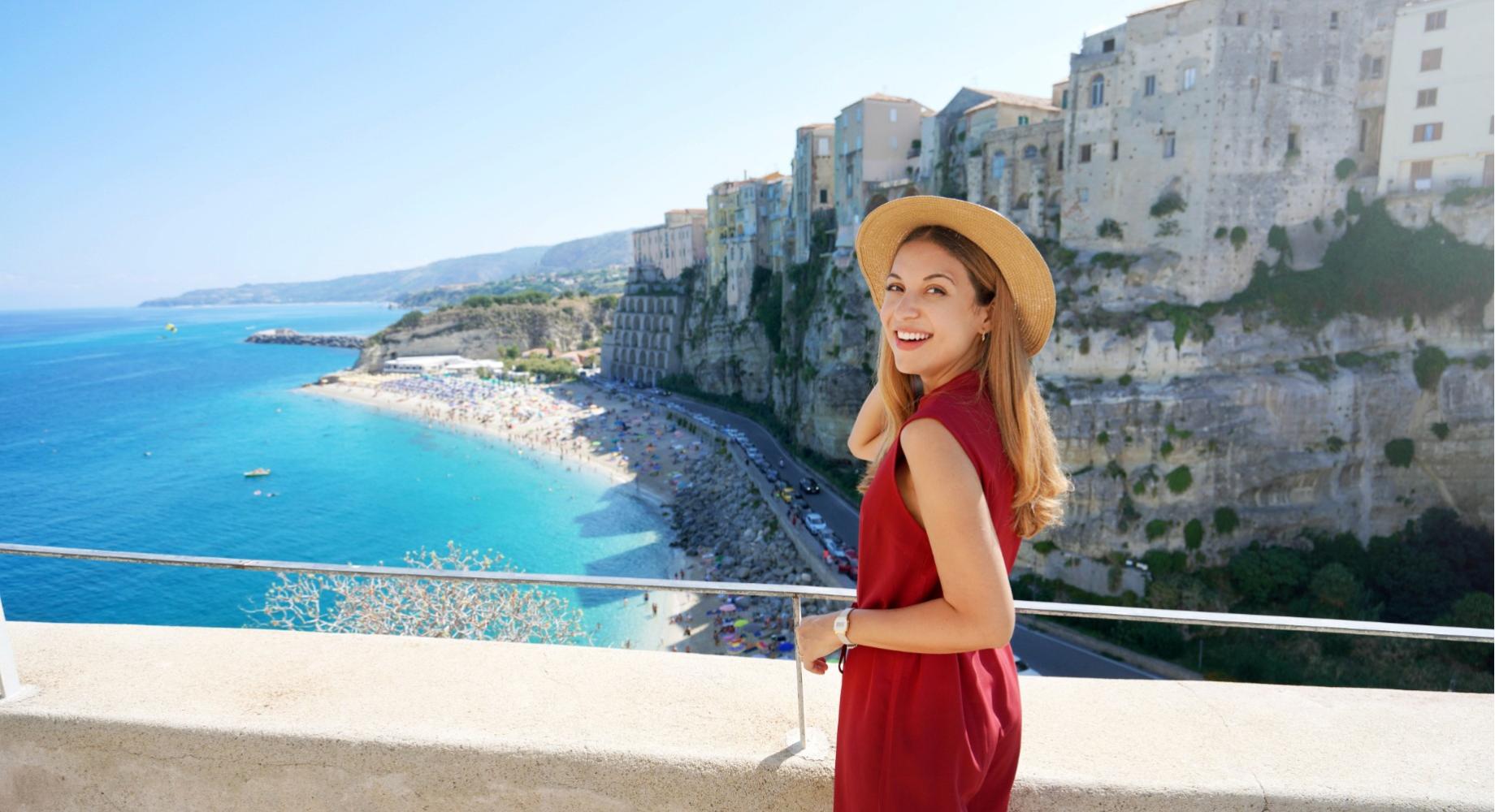 Donna sorridente con cappello, vista mare e scogliere, giornata soleggiata.