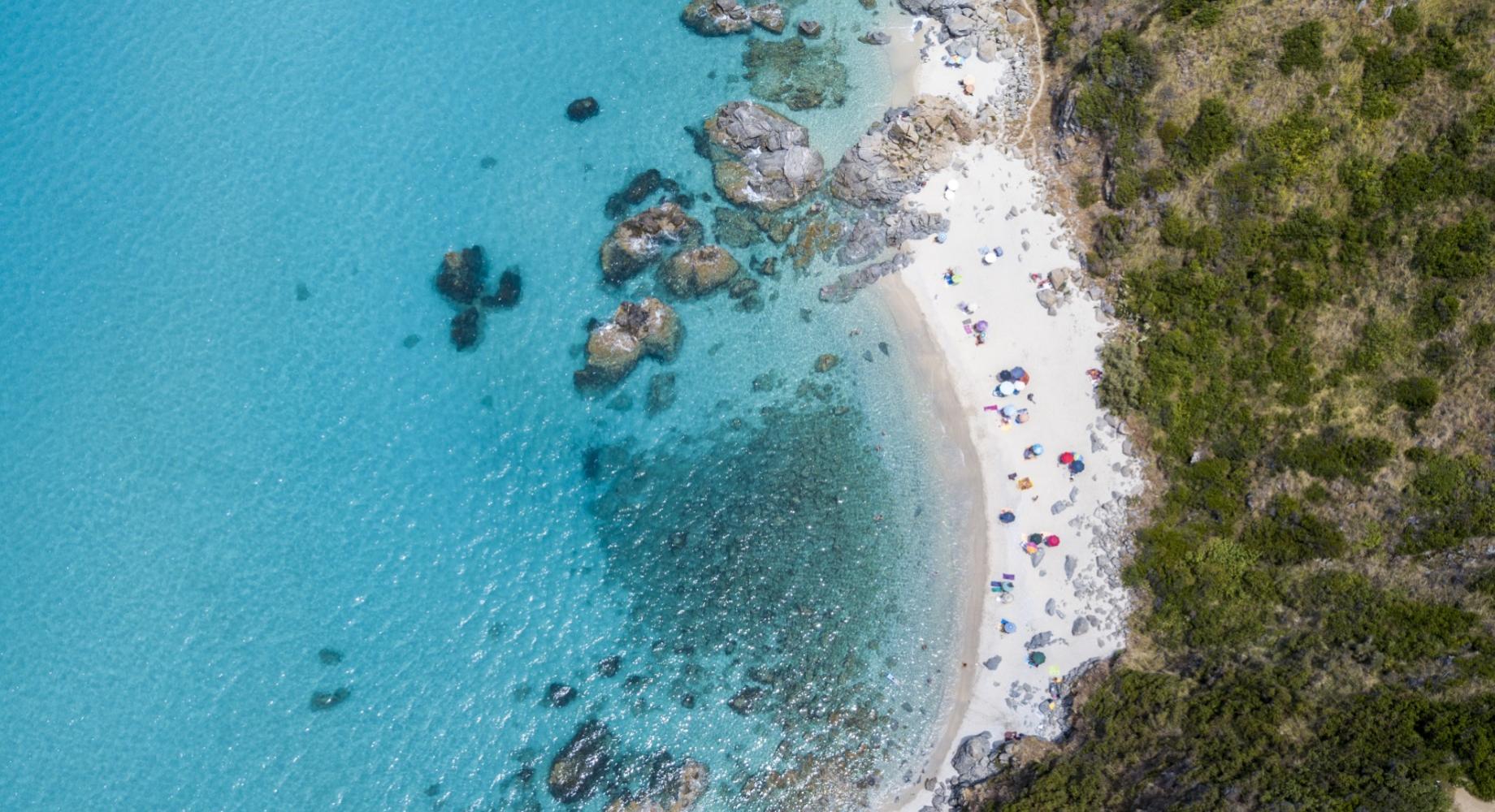 Spiaggia con mare cristallino, rocce e ombrelloni colorati vista dall'alto.