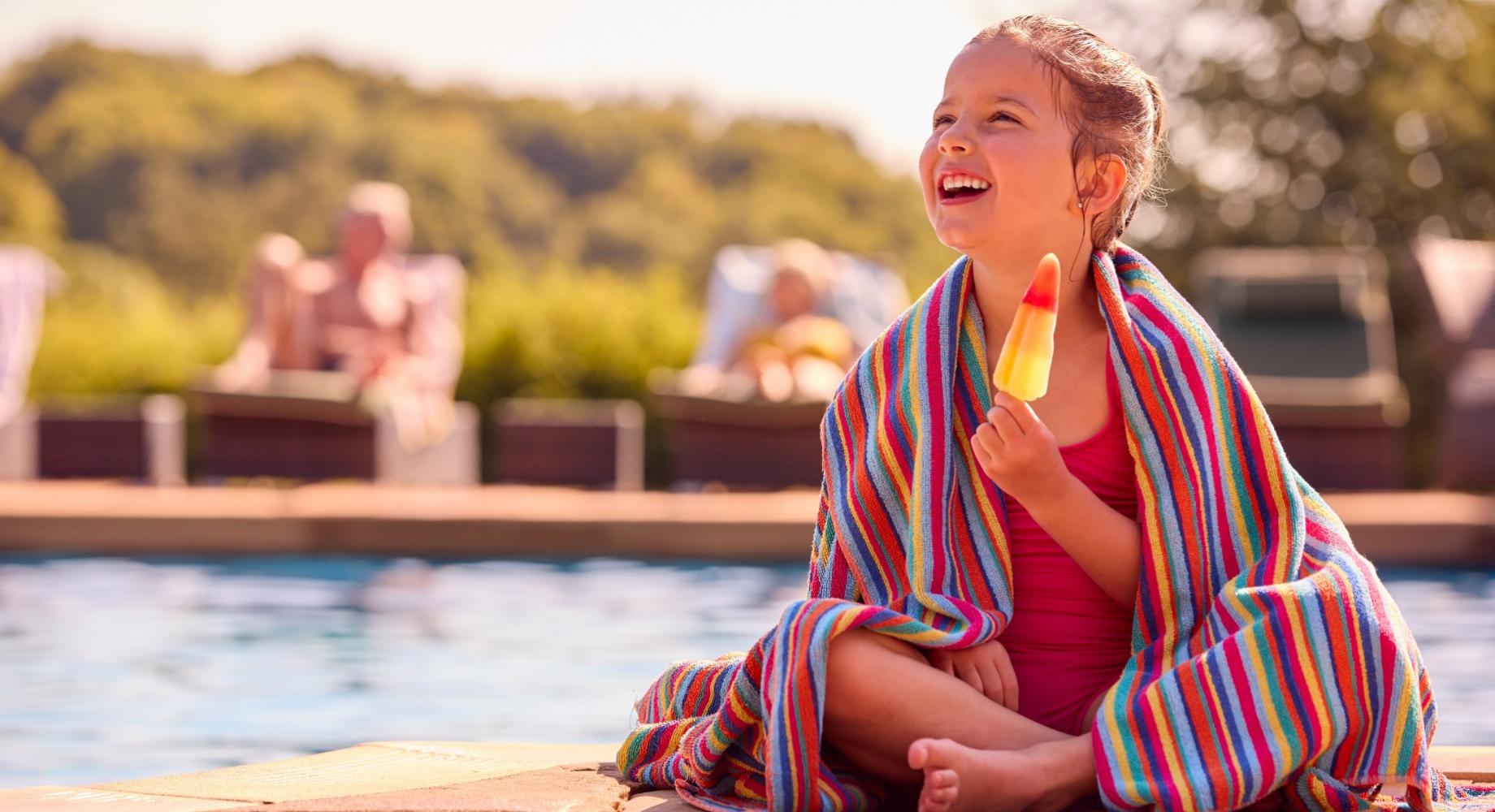 Bambina felice mangia un ghiacciolo vicino alla piscina, avvolta in un asciugamano colorato.