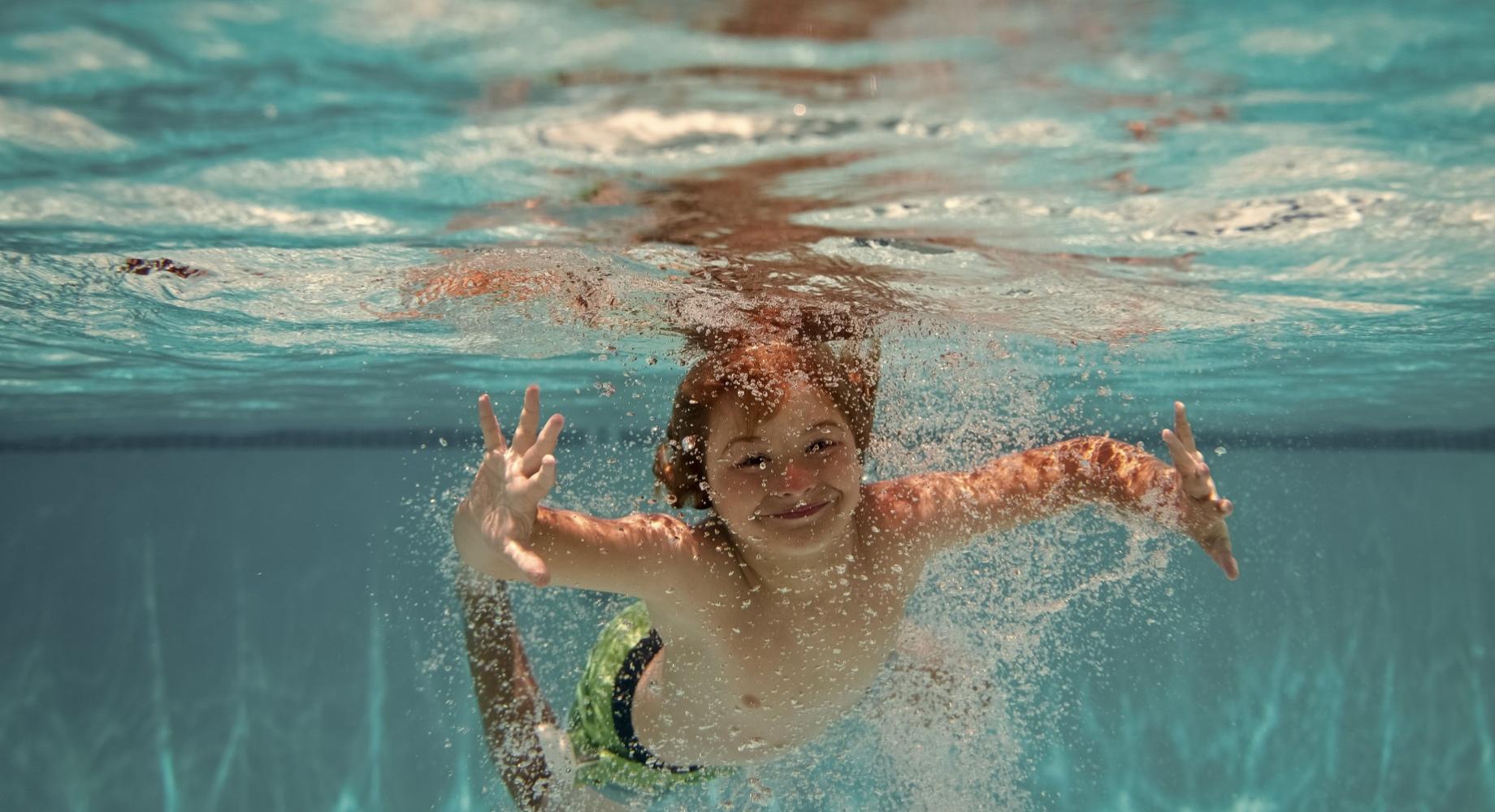 Bambino nuota sott'acqua in piscina, sorridente e circondato da bolle.