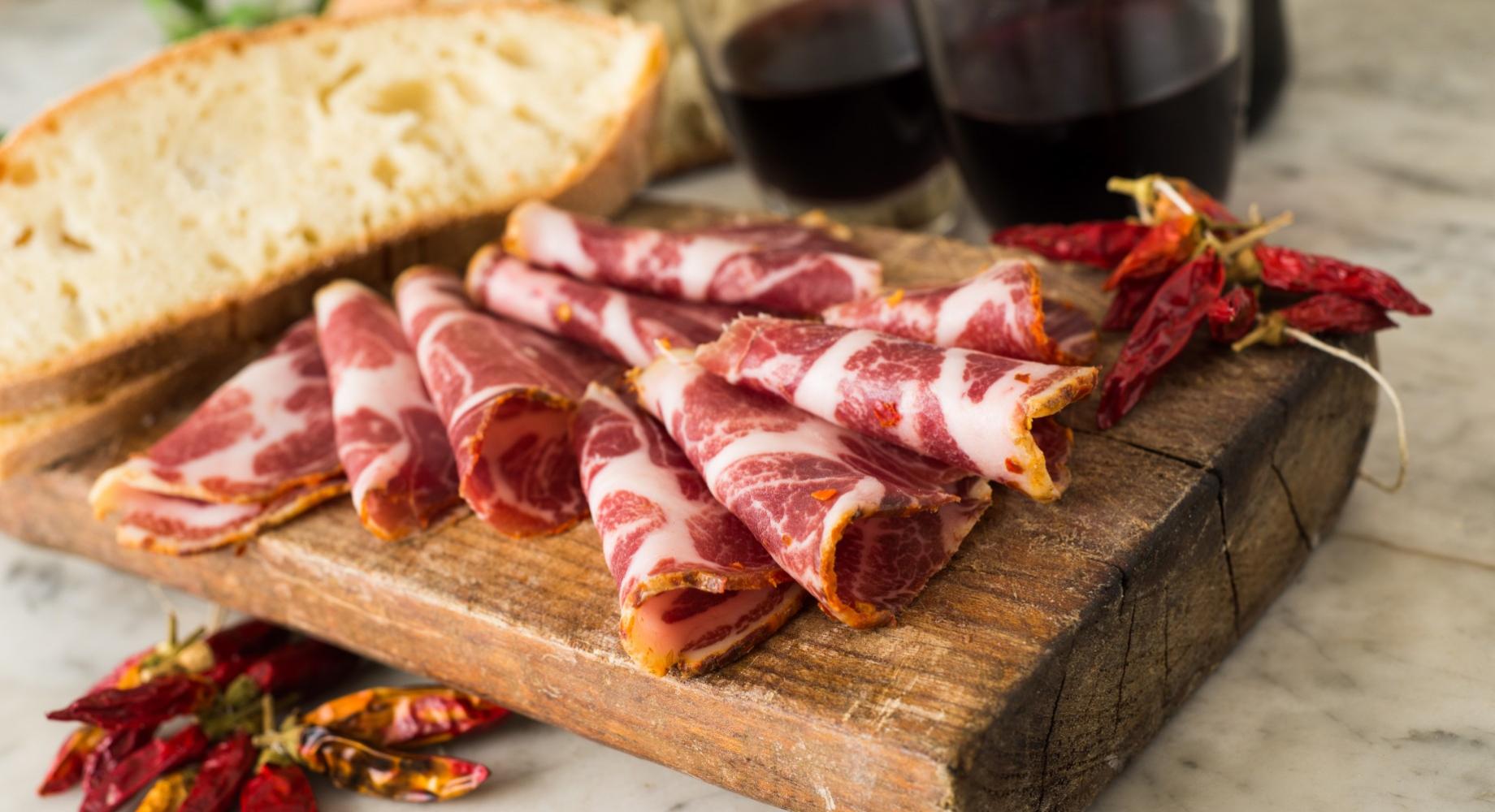 Salumi affettati su tagliere con pane, peperoncini e bicchieri di vino rosso.