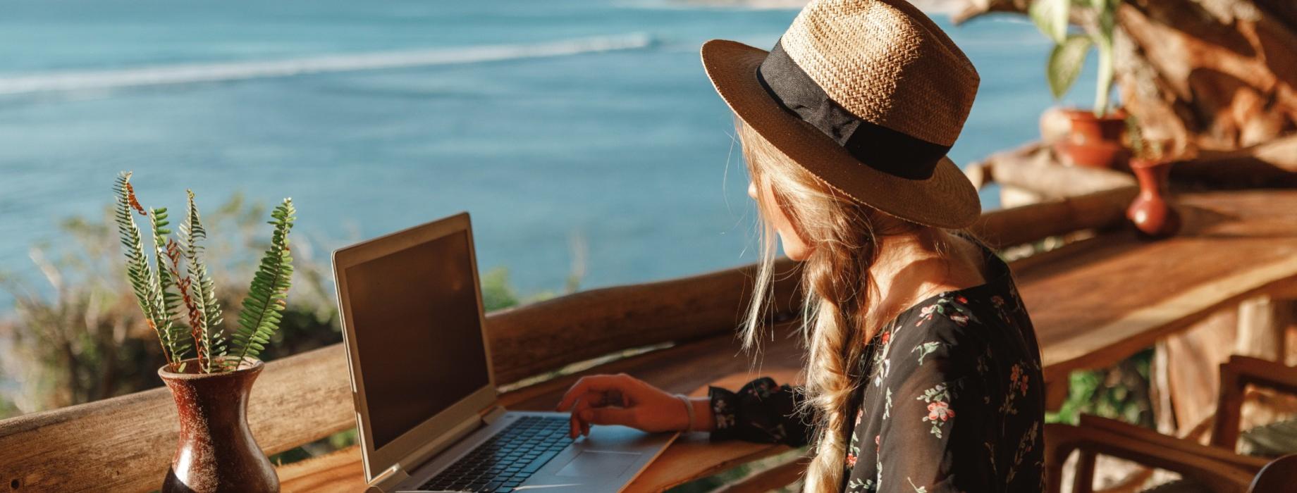 Donna con cappello lavora al laptop, vista mare, atmosfera rilassante.