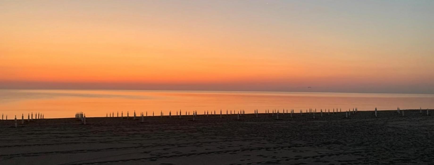 Tramonto sul mare con scogliere e spiaggia, cielo nuvoloso e barche lontane.