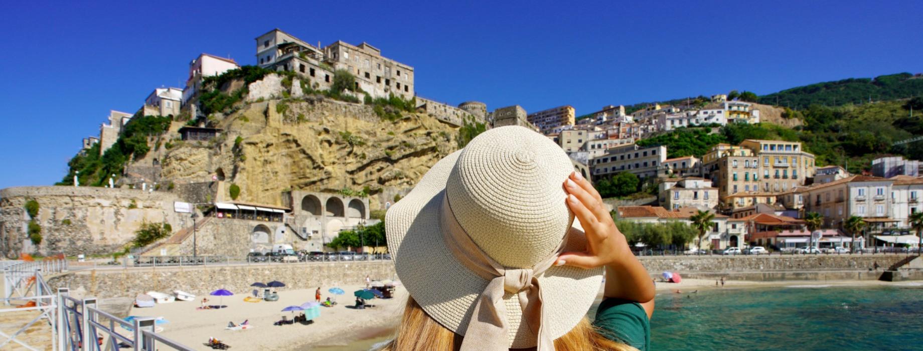 Donna con cappello ammira un borgo costiero arroccato su una scogliera.