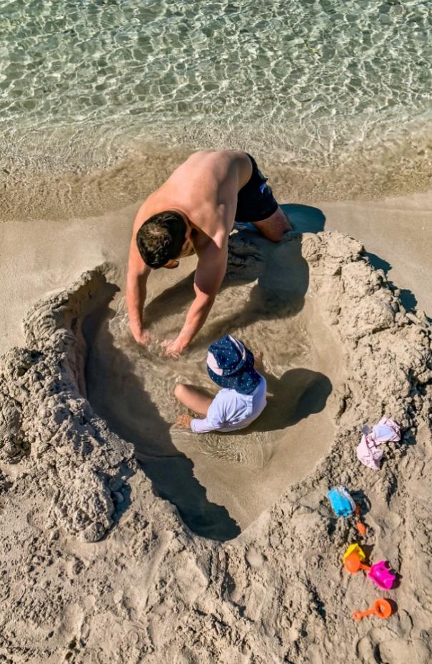 Uomo e bambino giocano in una buca sulla spiaggia con giocattoli colorati.